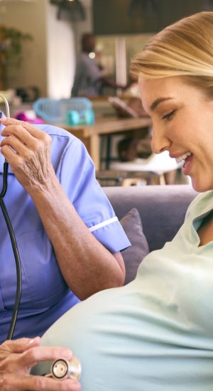 Senior Midwife With Stethoscope Visiting Pregnant Woman At Home With Family In Background